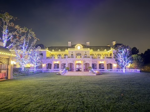 Large home viewed across its lawn with warm-white architectural lights and violet-lit trees