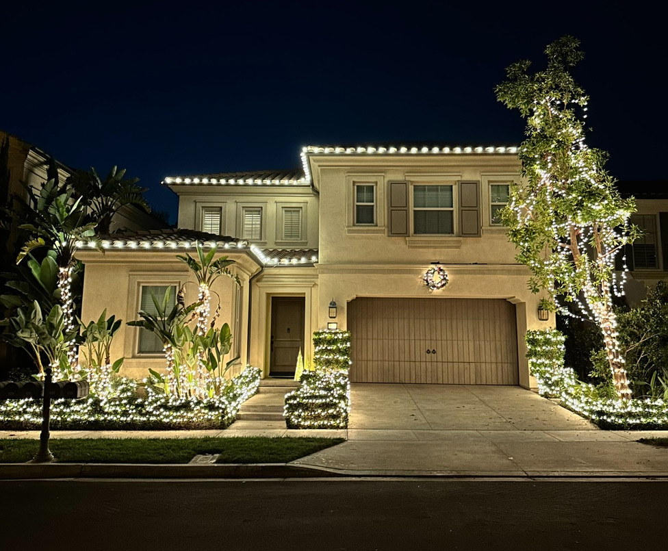 Two-story home outlined in warm white lights with illuminated trees and shrubs